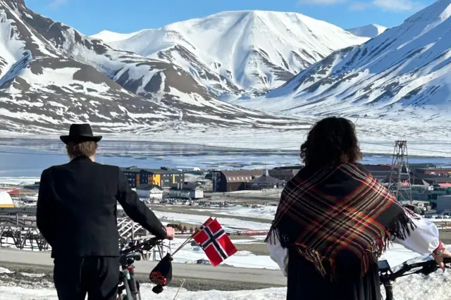 Norwegians wheeling their bikes on their national day in Svalbard, with water and icy peaks in the distance
