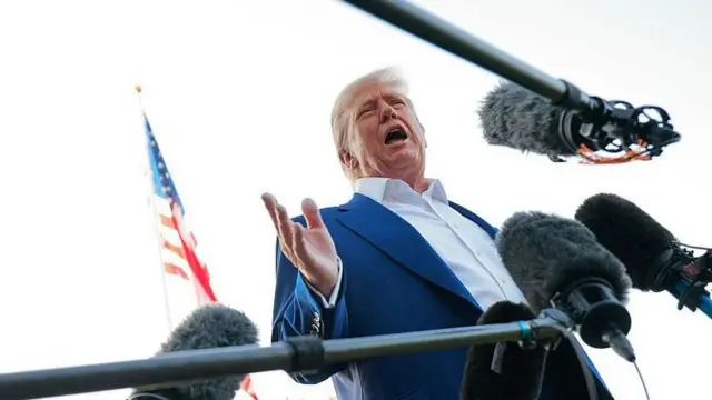 Hình ảnh Tổng thống Donald Trump mặc áo vest và áo sơ mi cổ mở, xung quanh là các micro, được chụp từ phía dưới.