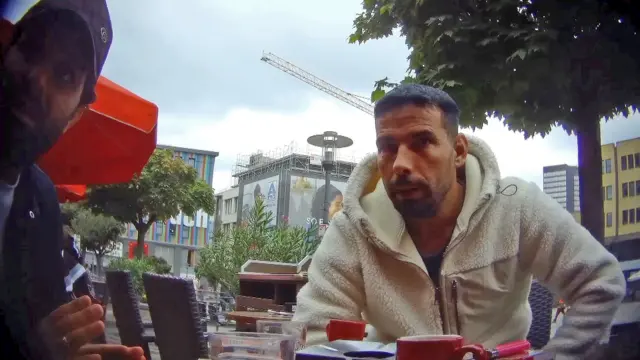 L'homme brun, barbu et aux cheveux courts, vêtu d'une veste polaire blanche, qui figurait sur les photos précédentes prises par le journaliste infiltré, discute dans un café situé sur un trottoir du centre d'Essen. Il y a des tasses de café sur la table, des gratte-ciel, des arbres et une grue en arrière-plan. Un deuxième homme - portant une casquette de baseball Gucci et une chemise bleue - avec une barbe et des cheveux foncés est visible à l'extrême gauche de la photo.