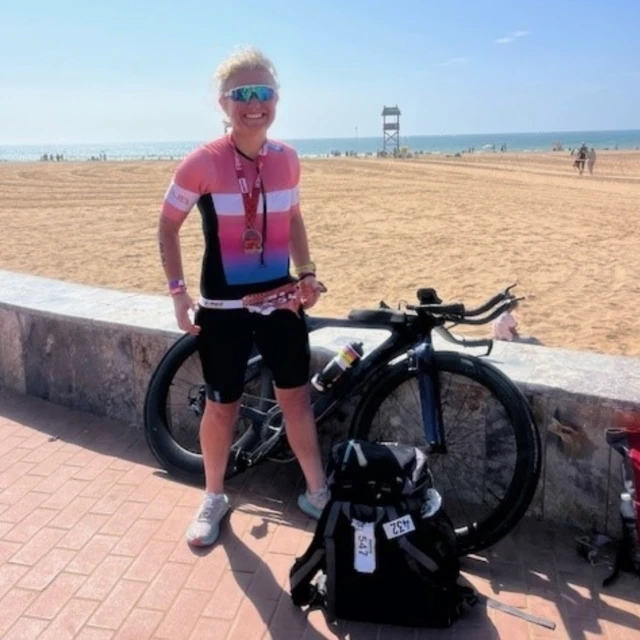 Una mujer rubia con el pelo recogido está de pie junto a una bicicleta con una playa de arena al fondo. Lleva un traje de ciclismo de licra rosa, blanco, azul y negro y una medalla colgada del cuello. Lleva gafas de sol de ciclismo azules reflectantes.
