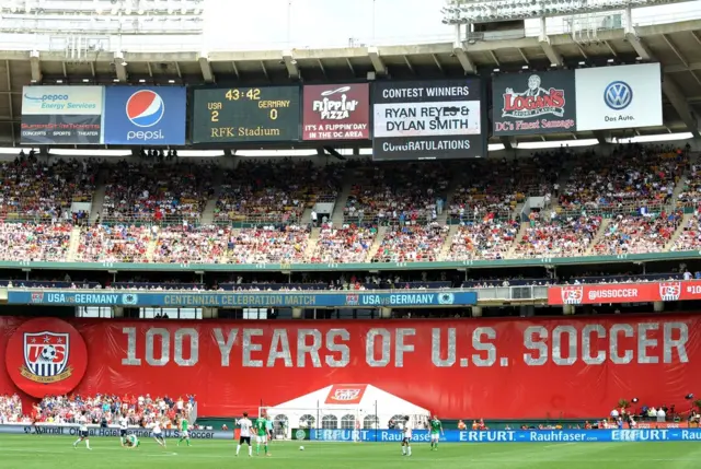 Los jugadores juegan en la cancha. Se ven las gradas del estadio y una pancarta gigante de fondo rojo que dice 100 years of U.S. Soccer. Hay una pantalla electrónica que muestra el tiempo y el marcador: 2 para USA y 0 para Alemania. Se ven publicidades de compañías.