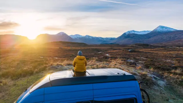 Sarı palto ve bere giymiş bir kişi, bir karavanın üzerinde oturmuş, ufuk çizgisinin hemen üzerinde yükselen güneşle birlikte dağ manzarasına bakıyor.
