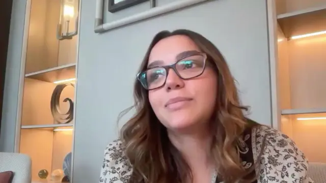 Uma mulher de cabelos castanhos ondulados, usando óculos de armação escura e uma blusa clara com estampa floral, sentada em uma poltrona. Ela olha levemente para o lado. Ao fundo, há prateleiras embutidas com iluminação quente, exibindo objetos decorativos como esculturas e esferas de vidro, além de uma luminária de parede com design moderno, criando um clima acolhedor e elegante.