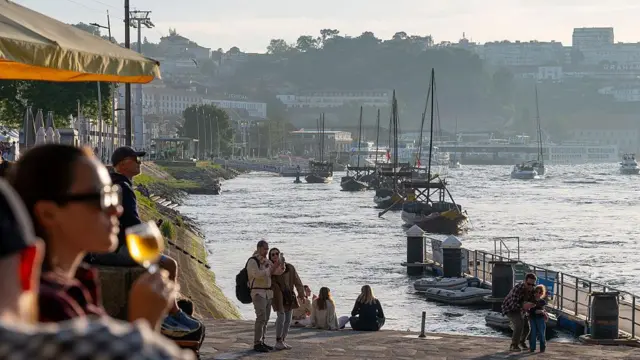 Zachód słońca w Porto na północy Portugalii. Ludzie siedzą nad brzegiem rzeki Douro. Jedna z kobiet, na pierwszym planie po lewej, trzyma kieliszek wina. Inni przechadzają się nabrzeżem i robią zdjęcia.