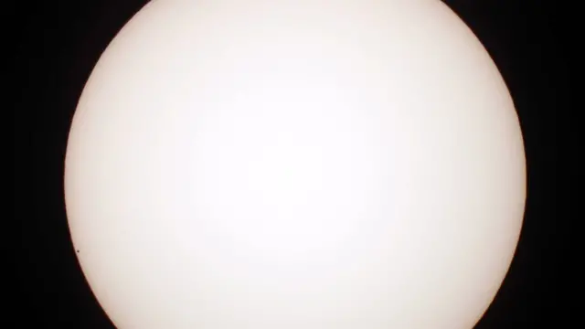 Una foto del Sol que lo toma como un gran disco blanco y en el extremo izquierdo a un diminuto punto negro que es Mercurio