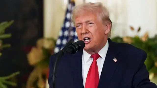 Президент Трамп в темном костюме и красном галстуке говорит в микрофон