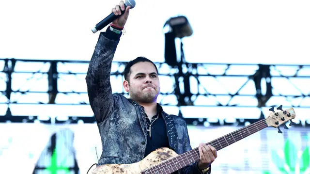 Ernesto Barajas, parado sobre un escenario, levanta su brazo derecho sosteniendo un micrófono y con una guitarra colgada.