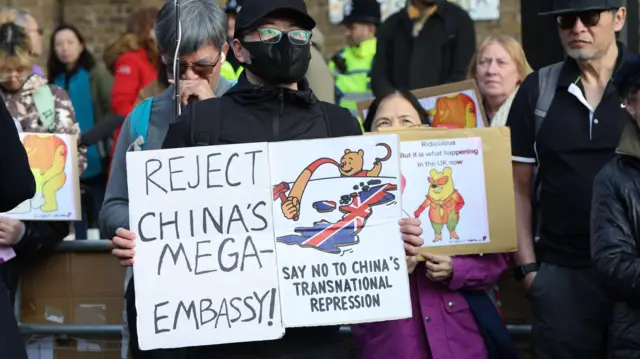 倫敦前皇家鑄幣廠外一名華人示威者舉起反對興建中國「超級大使館」的標語牌，顯示維尼熊伸長手臂，舉起共產黨黨徽上的鐵鎚，碰觸披上英國國旗的英國國土（17/1/2026）