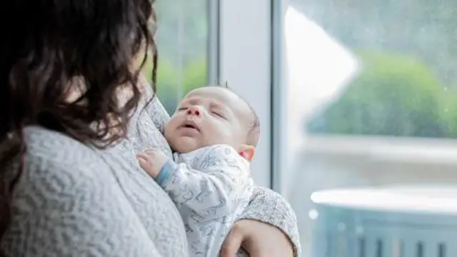 Молода дівчина із розпущеним темним волоссям стоїть біля вікна і тримає на руках немовля.