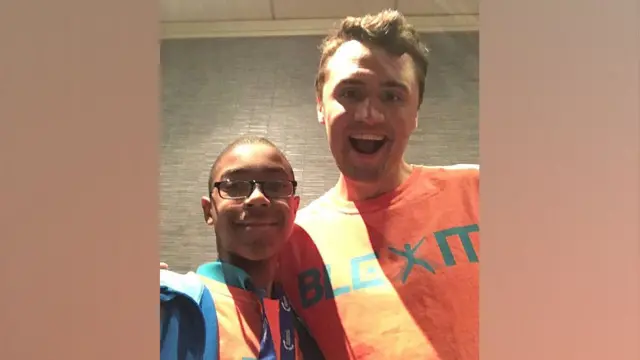 Chandler Crump, con gafas, una sudadera con capucha azul y una camiseta naranja brillante, sonríe en una foto estilo selfie con Charlie Kirk, quien lleva una camiseta naranja similar. 