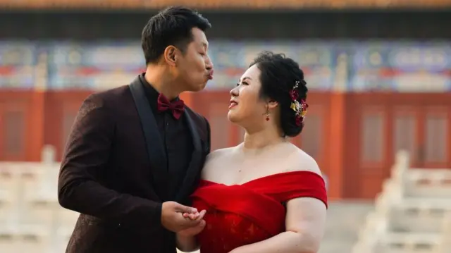 عکس عروس با لباس عروسی سرخ و داماد با دریشی سیاه که لبهای خود را برای بوسیدن عروس به صورتش نزدیک کرده است.