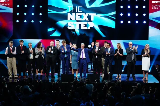 Nigel Farage sahnede, kalabalığa el sallıyor. Yanında alkış tutan çeşitli kadın ve erkekler var.