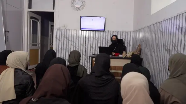 معلم که چادر و ماسک صورت سیاه دارد، پشت میز نشسته و به کامپیوتر نگاه می‌کند. شاگردان به زمین نشسته‌اند. 