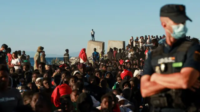 Migranci stoją w kolejce do punktu Czerwonego Krzyża w Ceucie. Na pierwszym planie funkcjonariusz służb bezpieczeństwa w masce ochronnej. Na dalszym planie morze i widoczne w oddali sylwetki wzgórz. Źródło: Reuters / Violeta Santos Moura