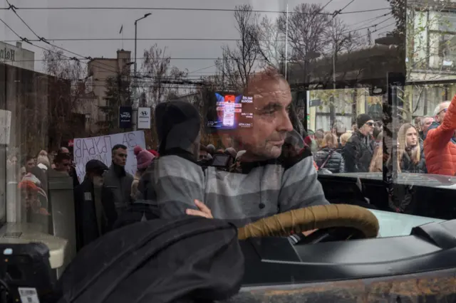 یک راننده اتوبوس از پشت فرمان، به مردمی نگاه میکند که برای ادای احترام به پانزده قربانی، خیابانی را در بلگراد به مدت پانزده دقیقه مسدود کردهاند