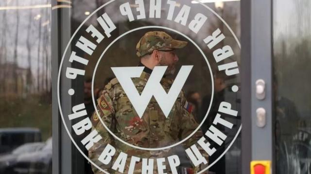 Homem em traje militar atrá de vidro com logotipo do grupo Wagner