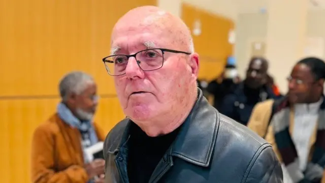 Jenerali Christian Quesnot yambaye indorerwamo n'ikoti ry'uruhu ry'umukara, inyuma ye hahagaze abandi bantu 