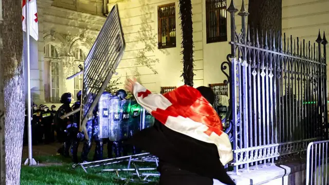 Человек с грузинским флагом у сломанного забора, за забором стоит полиция.