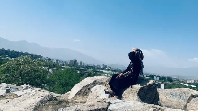 نمای جانبی زن با حجاب سیاه و صندل نشسته روی صخره ها با ساختمان‌ها و کوه‌ها در پس زمینه
