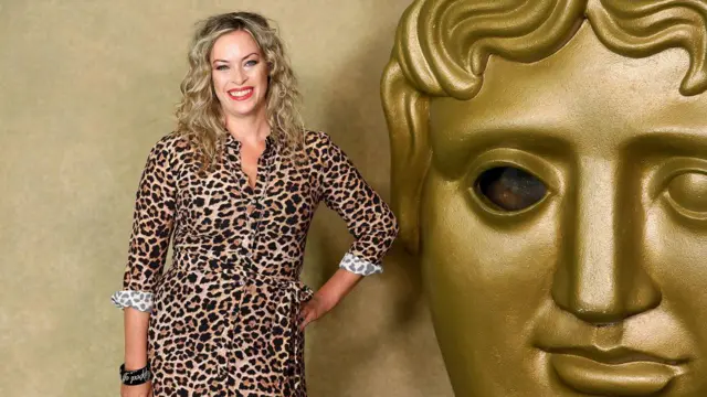 Sharon - kobieta o falowanych blond włosach - stoi obok dużego napisu BAFTA. Ma na sobie sukienkę w panterkę. Uśmiecha się i trzyma dłoń na biodrze.