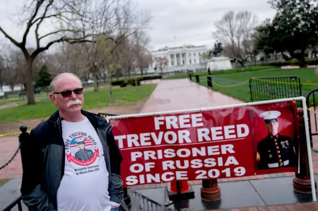 Чоловік із сивим волоссям в темних окулярах біля плаката "Звільніть Тревора Ріда. Вʼязень Росії з 2019 року