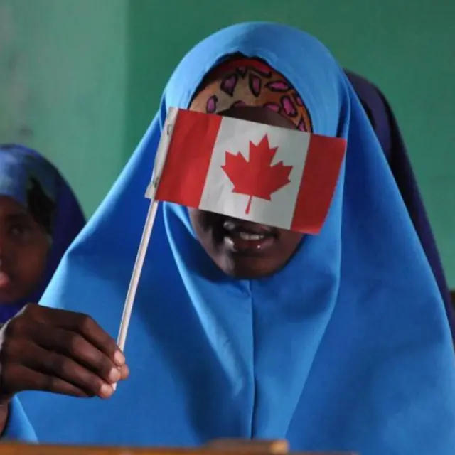 A school girl in blue hijab waves a tiny Canada flag across her face 
