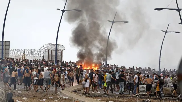 Un fuego y el humo que produce se elevan por el aire en medio de cientos de personas junto a la frontera terrestre en Marruecos y España en Ceuta.