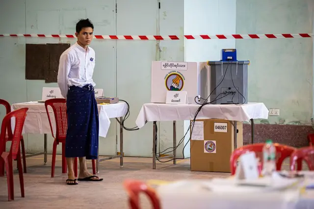 မဲပေးစက်နားမှာရပ်နေတဲ့ ရှပ်အဖြူ၊ ပုဆိုးအပြာဝတ်ထားတဲ့ မဲရုံဝန်ထမ်းတစ်ဦး 