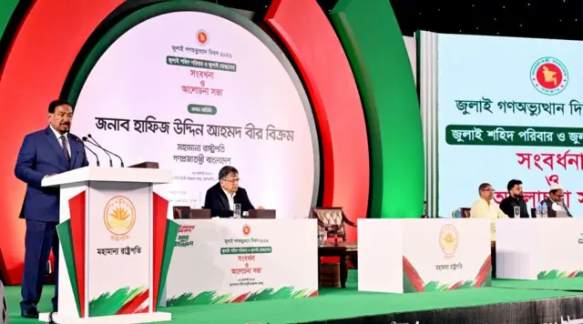 হাফিজ উদ্দিন আহমেদ ডায়াসে দাঁড়িয়ে বক্তব্য দিচ্ছেন। পাশেই টেবিলের পেছনে বসে আছেন সালাহউদ্দিন আহমেদ