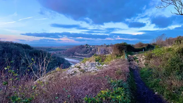 Un cielo azul con dramáticas nubes azules y naranjas se extiende sobre la garganta del Avon. Los árboles y setos son muy escasos, con un toque de verde visible entre las ramas y las rocas. Hay una luz casi púrpura en las ramas. El río, con marea muy baja, se puede ver fluyendo por la garganta.
