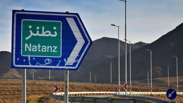 Uma placa de trânsito azul e verde que diz “Natanz” no Irã fica ao lado de uma rodovia, com uma cordilheira ao fundo