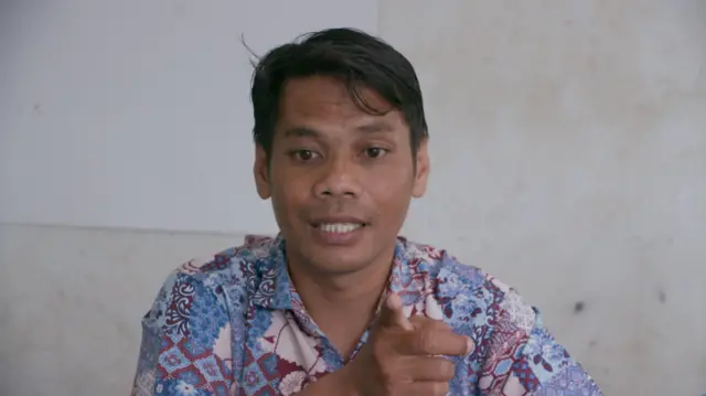 Zurhan Santi mengenakan baju batik biru keunguan, tangannya menunjuk ke depan.