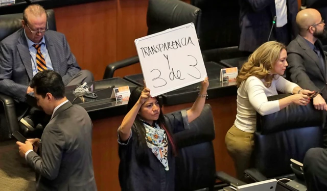 Indira Kempis 'Transparencia y 3 de 3' kelimelerinin yazılı olduğu bir tahtayı senatoda elinde tutuyor - etrafındaki siyasetçiler telefonları ile ilgileniyor, konuşuyor ya da bir şeyler okuyorlar.