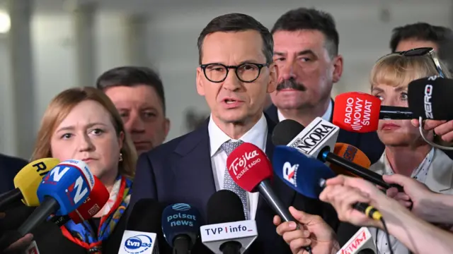 Mężczyzna z ciemnymi, krótkimi włosami ,w czarnych okularach, białej koszuli i w niebieskiej marynarce stoi w otoczeniu pięciu osób, przed nimi znajdują się mikrofony prasy