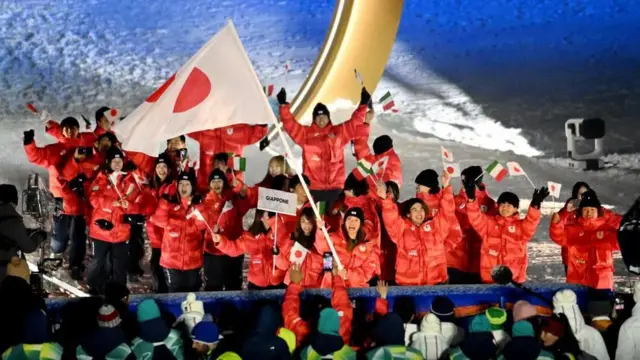 そろいの赤い上着と黒っぽい帽子を身に着けた選手たちが、日本とイタリアの国旗を持って両手を上げている