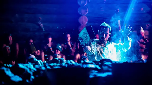 Mężczyzna w białej koszuli polewa rozgrzane kamienie wodą. W tle widoczni są ludzie siedzący w saunie.