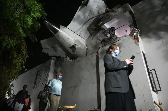 Personas caminando con mascarillas pasan por un edificio dañado que tiene en el techo restos del avión