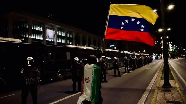 تظاهرکننده‌ای که پرچم ایران را به تن دارد، در حالی که از میان پلیس ضدشورش در نزدیکی سفارت ایالات متحده در آتن یونان عبور می‌کند، پرچم ونزوئلا را تکان می‌دهد.