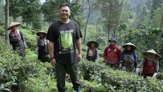 Rizal Firdaus, mengenakan kaos berwarna hitam bergambar daun teh, dengan tulisan tea-shirt, berfoto bersama seorang petani dan lima orang perempuan pemetik teh di kebun.