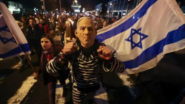 تصویر یک مخالف نتانیاهو را با پرچم اسرائیل نشان میدهد. ده‌ها نفر دیگر از معترضان در پشت او دیده می‌شوند.