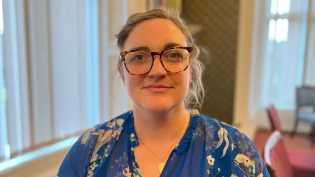 Una persona con una blusa azul estampada con flores está sentada en el interior de una habitación con persianas verticales altas, papel tapiz estampado y sillones tapizados. Al fondo se ven un radiador y obras de arte enmarcadas.