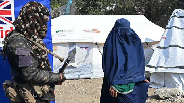 یوه ښځه چې شنه چادري/برقه یې پر سر ده، روانه ده او څنګ ته د طالبانو حکومت یو سرتېری وسله په لاس ولاړ دی چې په برګ دسمال یې سر او مخ پوښلي دي.