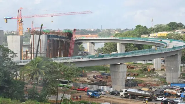 Le chantier du pont de Yopougon à Abidjan en mars 2023 montrant des grues, des piliers en béton et une passerelle courbe surélevée.
