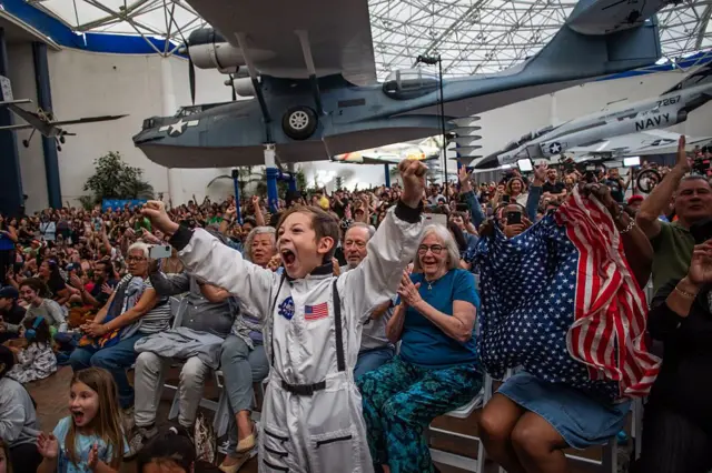 Un niño vestido con un disfraz de astronauta anima junto a una mujer que ondea una bandera mientras ven una retransmisión en directo del regreso de los tripulantes del Artemis II a la Tierra en el Museo del Aire y el Espacio de San Diego
