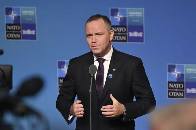 Навроцький в діловому костюмі виступає перед журналістами біля стенда саміту НАТО в Анкарі. На тлі видно логотипи НАТО та напис «Ankara Summit 2026», попереду — мікрофон і телекамери.
