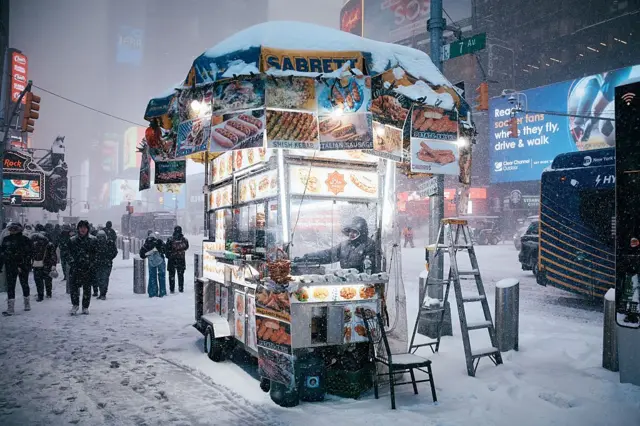 رجل يرتدي سُترة بكُمَّين، طويلة، تغطِّي النصف الأعلى من جسم، ويغطي رأسه، وذلك أثناء عمله في عربة طعام خلال عاصفة ثلجية بمدينة نيويورك، وتظهر الثلوج بينما تكسو أرض الشوارع.