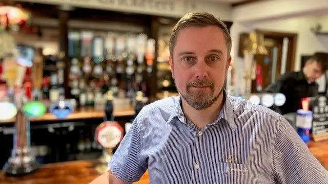 Matthew Allum, con barba y camisa azul con el cuello abierto, se encuentra en su bar.