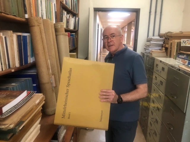 Professor na sala dele, em pé, segurando grande envelope e olhando em direção à câmera com olhar sério