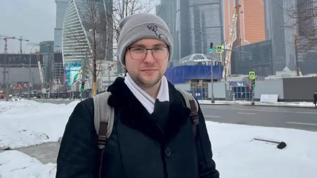 A kép előterében egy férfi látható szürke kötött sapkában, szemüvegben, fekete kabátban és fehér sálban. Hátizsákot visel. A járdát és az út szélét hó borítja. A háttérben széles utca húzódik közlekedési lámpákkal és gyalogátkelőhely-jelző táblákkal. Távolabb modern, üveg homlokzatú magas épületek és daruk láthatók. Lombtalan fák szegélyezik az utcát, és az út közelében iépítkezés és kordonok láthatók.