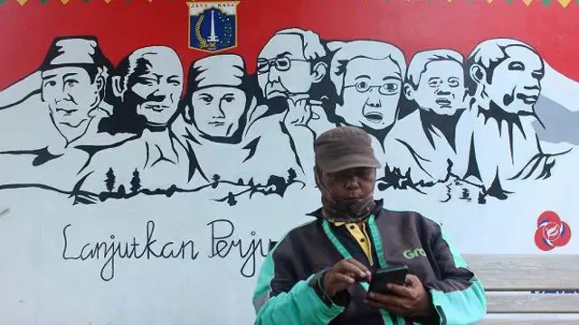 Pengemudi ojek dalam jaringan duduk di depan mural bertema Pemilu 2024 di Jalan Perjuangan, Jakarta Barat, Senin (20/11/2023).